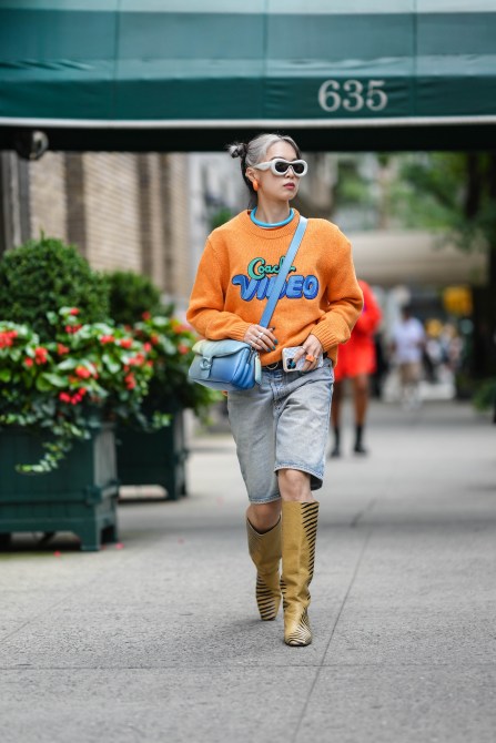 Woman wears white latte puffy leather sunglasses from Loewe, a blue necklace, orange earrings, an orange with embroidered green and blue pattern pullover, a blue to beige gradient print pattern puffy leather crossbody bag from Coach, blue faded denim shorts, beige shiny leather with black striped print pattern heels knees boots / high boots, rings , outside Coach, during New York Fashion Week