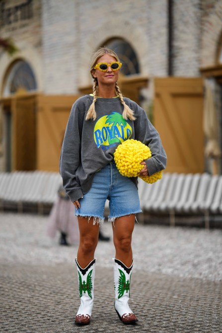 Woman wears yellow flower sunglasses, a yellow t-shirt, a gray with yellow / blue / green print pattern sweater, a yellow large fluffy embossed fringed handbag, blue faded denim ripped knees shorts, white leather with green / brown embroidered pattern block heels knees boots , outside Munthe, during the Copenhagen Fashion Week