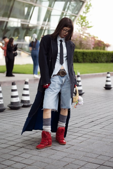 A guest wears long grey socks, red boots, blue denim shorts, white shirt, black tie, red gloves, beige bag with attached accessories and long black coat on day 2 of Visa Fashion Week Almaty 2024 on May 02, 2024 in Almaty, Kazakhstan.