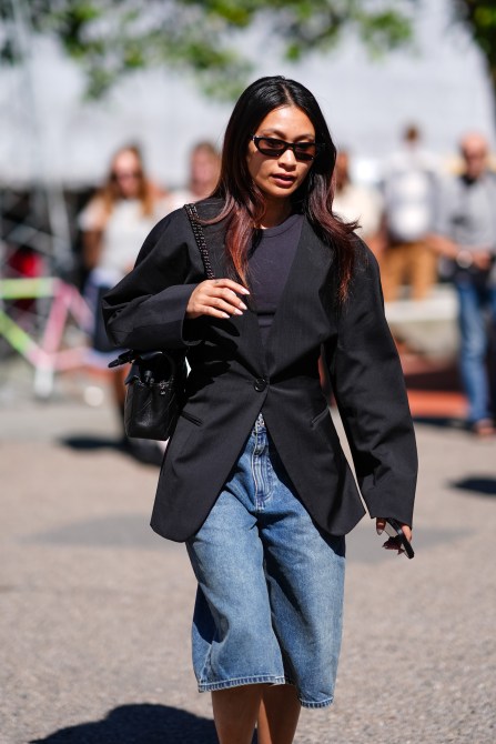 COPENHAGEN, DENMARK - AUGUST 6: A guest wears black sunglasses, black shirt, black tailored blazer jacket, shiny black leather bag, light blue denim jean jorts, outside Joao Maraschin, during the Copenhagen Fashion Week Spring/Summer 2024-2025 on August 6, 2024 in Copenhagen, Denmark.