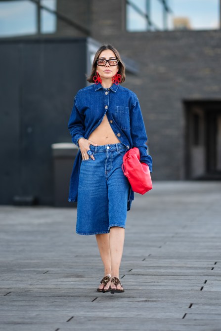 COPENHAGEN, DENMARK - AUGUST 5: A guest wears black sunglasses with orange lenses, bright red floral earrings, dark navy blue denim jean half button long sleeve dress, bright red drawstring bag, a blue ring, light navy blue denim jean jorts, dark brown sandals, outside Forza Collective, during the Copenhagen Fashion Week Spring/Summer 2024-2025 on August 5, 2024 in Copenhagen, Denmark.