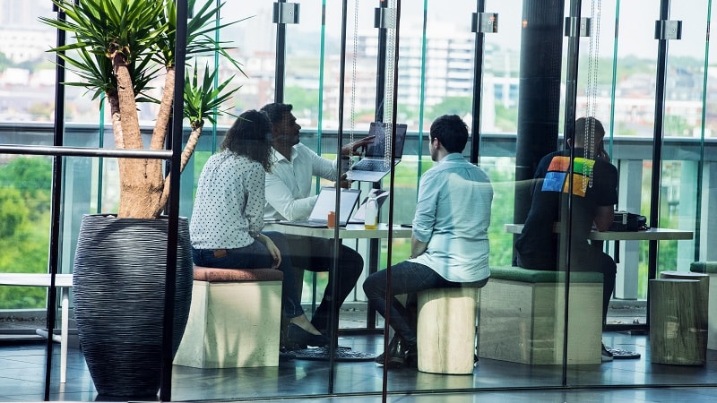 Cuatro personas colaborando en una oficina moderna con grandes ventanas y vistas a la ciudad