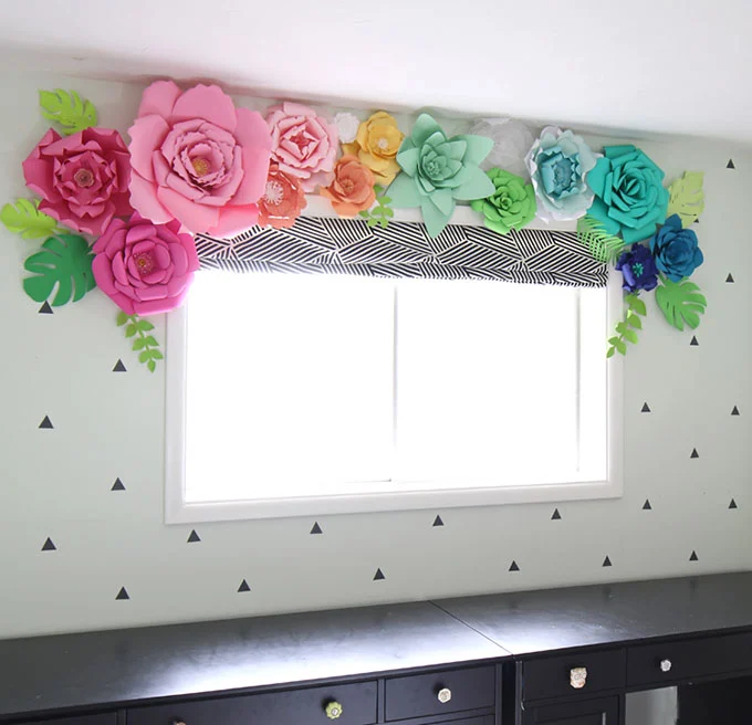 The top of a window decorated with various sized paper flowers in a rainbow of colors.