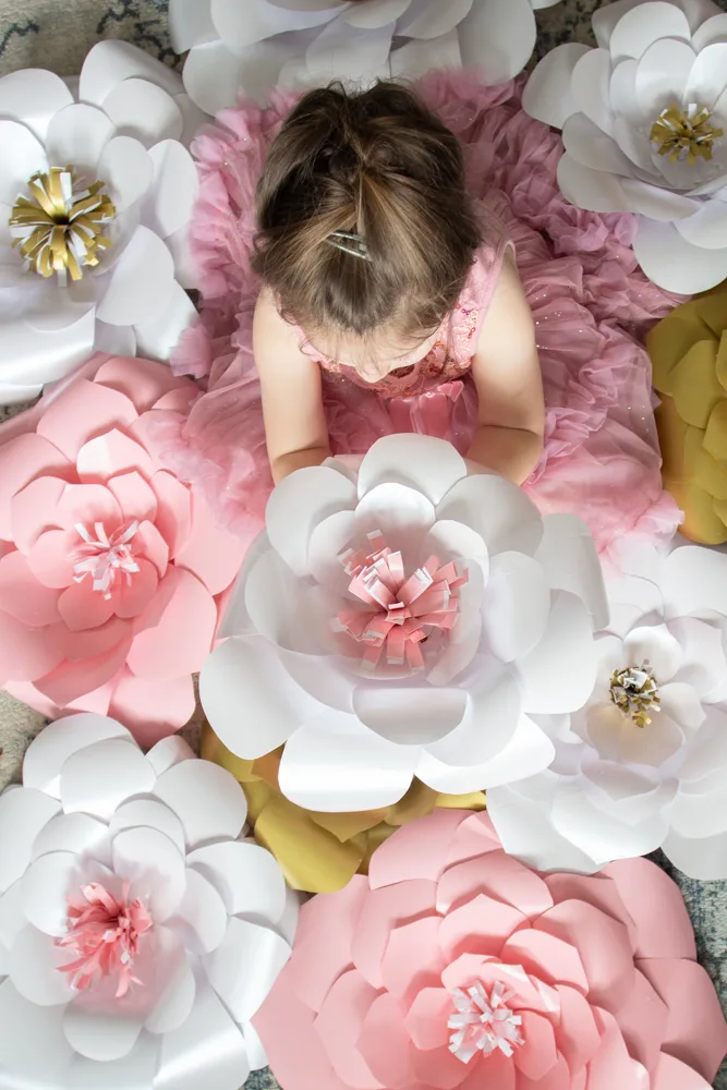 Small child in a pink dress holding up a large white paper flower surrounded by additional pink, white and gold giant flowers.