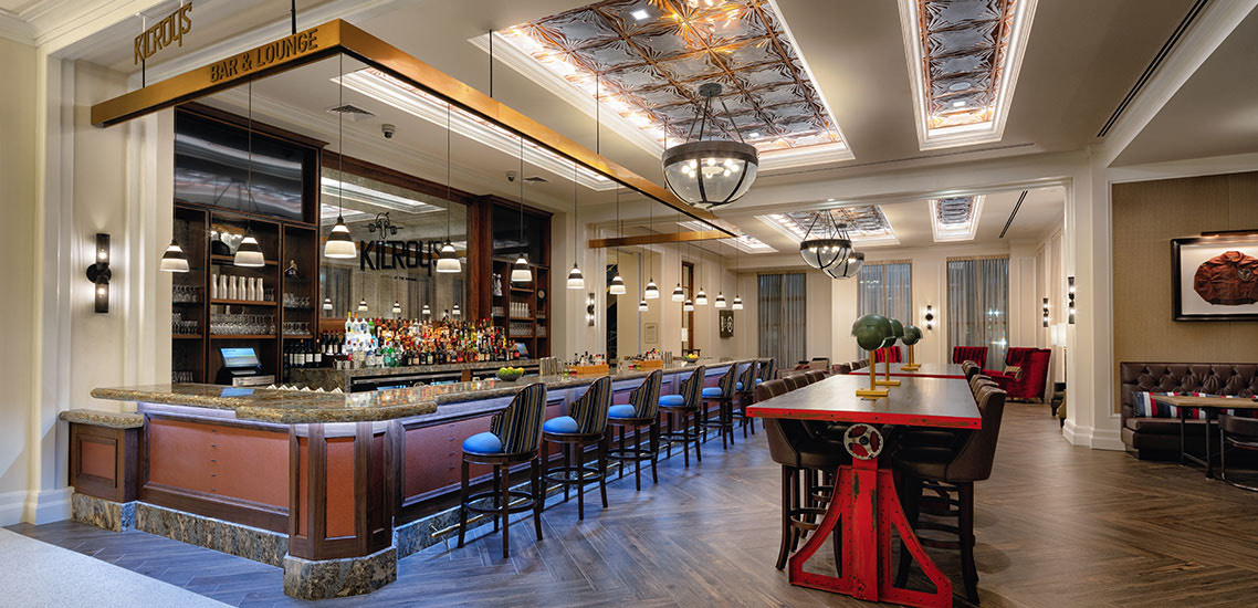 View of a room with a bar with several blue and black stools next to two tables 