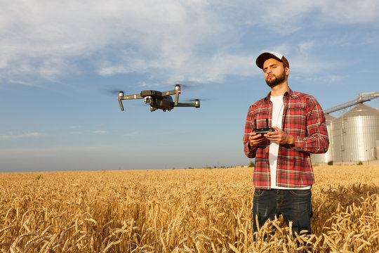 Drone hovers in front of farmer with remote controller in hands near grain elevator. Quadcopter flies near pilot. Agronomist taking aerial photos and videos in a wheat field.