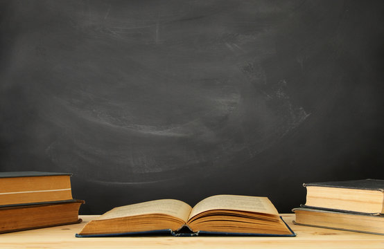 stack of books over wooden desk in front of blackboard.