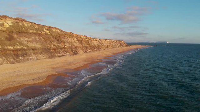 An aerial backward footage of the Hengistbury Head cliff with beautiful sandy beach and crystal blue water under a majestic blue sky and white clouds