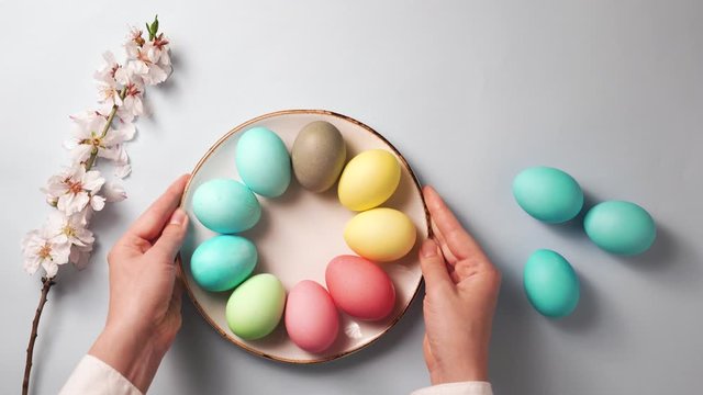 hands puts a plate with colorful eggs on an easter decorated table. branch with flowers and on a blue background. spring holidays concept. top view, 4K.