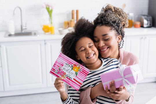 excited african american woman embracing happy daughter holding gift box and happy mothers day card