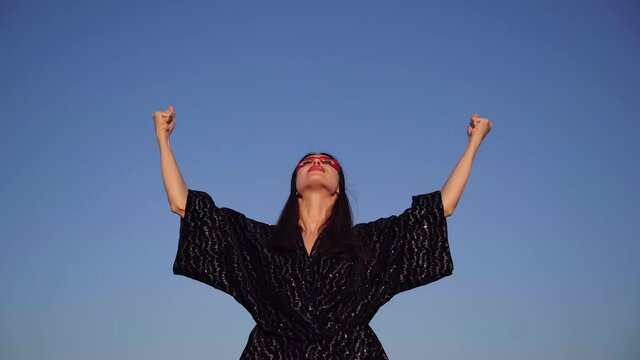 Brunette superhero girl in black dress and red face mask spreading hands with clenched fists and looking up on sky background. Female power, protest, women rights, activism concept. 4k video footage