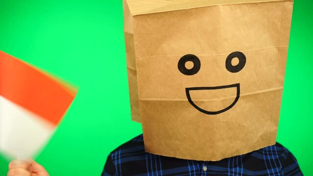 Portrait of man with paper bag on head waving Indonesian flag with smiling face against green background.