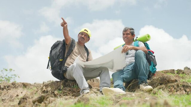 Middle aged Traveler friends checking route map for direction while sitting on top of hill during trekking - concept of recreation, exploration and journey