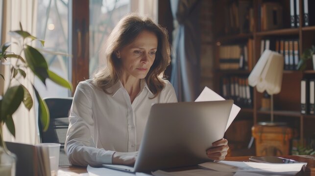 Busy 40 years old business woman working in office checking documents. Mid aged businesswoman accounting manager executive or lawyer using laptop reading paper file financial report, tax invoice.