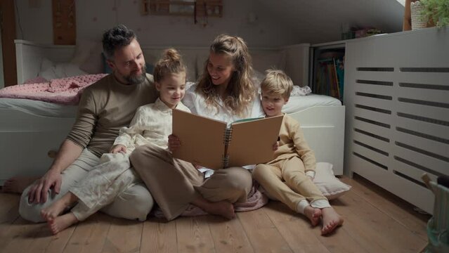 Beautiful family sitting in kids room on floor, looking at old family photos in photoalbum. Calm hygge family moment.