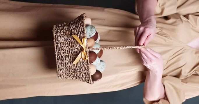 many chicken eggs in a basket in a woman’s hand for Easter painted in brown, blue and marble colors
