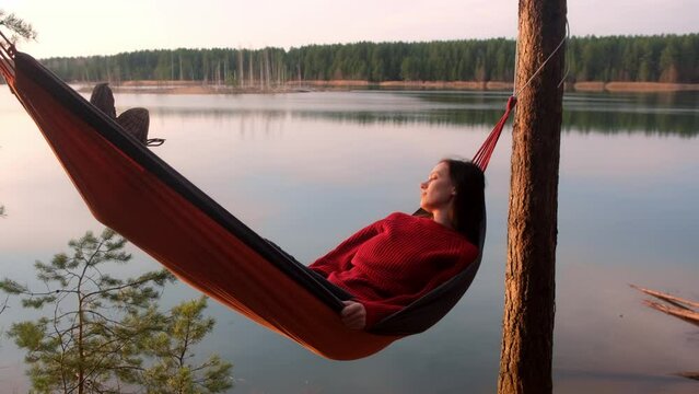 Relaxed woman lying in hammock in forest enjoy admiring lake nature at sunset. Resting relaxed young female in red sweater enjoying time in camping in dark woodland. Tourist traveler, camper outdoors.