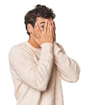 Young Hispanic man in studio blink through fingers frightened and nervous.