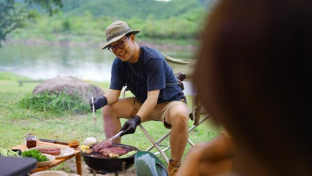 Group of Happy Asian man and woman enjoy and fun outdoor lifestyle travel nature camping in forest on summer vacation. People friends having dinner eating bbq grill and drinking beer together at night