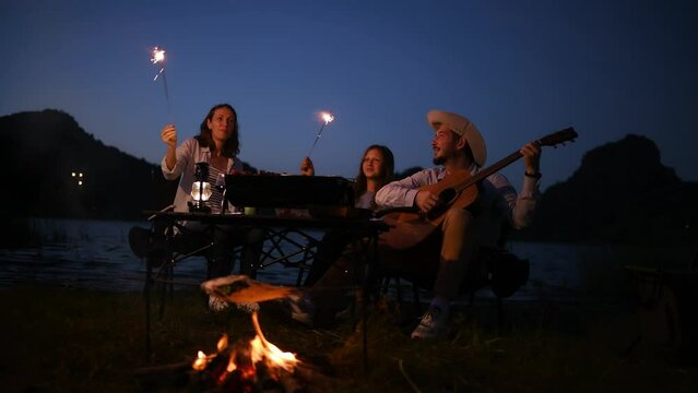 Group of travellers camping with friends they join drinking ,Bar-B-Q and playing guitar together, playing firework camping near nature lake with mountain background is holiday summer camping concept.