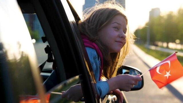 Close up of little joyful girl in vehicle window holding Turkish flag in hands. Beautiful child looking out of car window going on family trip. Small cute kid patriot with flag in car