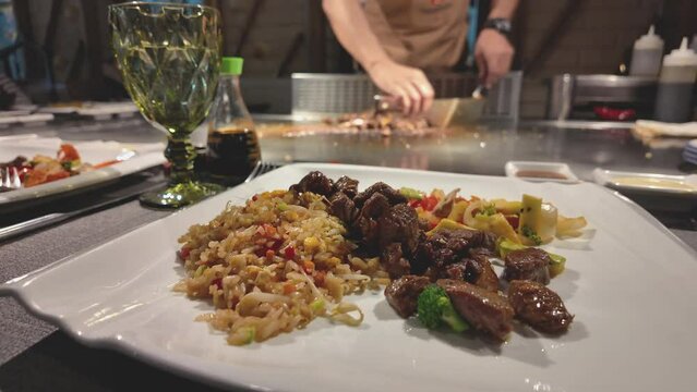 fried rice with vegetables and fried pieces of marbled beef with sliced fried champignons on a plate. In an out-of-focus background, a chef fries vegetables with rice on a teppanyaki frying table.