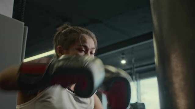 Low angle medium close-up of young female athlete in white tank top and boxing gloves practicing on punch bag at gym, delivering energetic strong blows in fast-paced routine