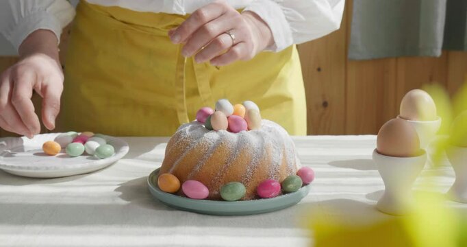 Female hands decorating Easter cake with small colorful chocolate eggs and daisy flowers. Anonymous woman making traditional easter cake or sweet bread with topping. Easter treat.