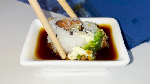 A close up view of a sushi roll being held with chopsticks and dipped into a mixture of soy sauce and wasabi sauce. Chinese food concept.