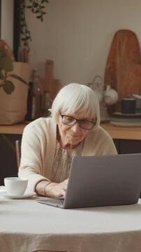 Vertical shot of elderly lady surfing internet on laptop while sitting at kitchen table with cup of tea