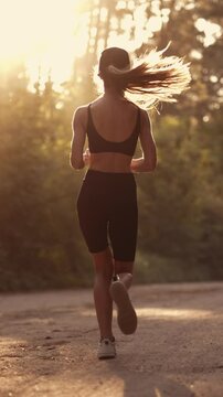 A young sporty woman runner is listening to music in earphones and training. Slim fitness girl is jogging outdoor. Concept of workouts running and healthy sport lifestyle. Vertical video footage