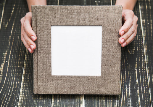 Closeup top view of female hands holding brown canvas photo book with empty white space in middle for text or photography. Album isolated on brown wooden background. Horizontal image with copyspace.