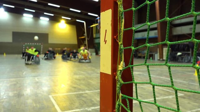 A close up view of a net in a sports hall. Disabled men in wheelchairs play rugby in the background and come closer to the net.