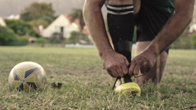 Closeup of a rugby player tying shoes on field with a rugby ball on the side. Rugby player adjusting his shoe laces on ground.
