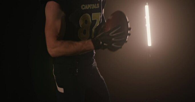 Muscular male rugby player in sportswear and helmet jumping with ball in dark studio