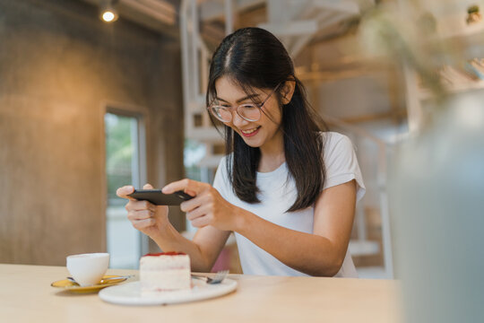 Blogger Asian friendly women influencer eat cake at night cafe. Beautiful young lady happy relax fun using technology mobile phone taking a photo of her food upload in social media at college campus.