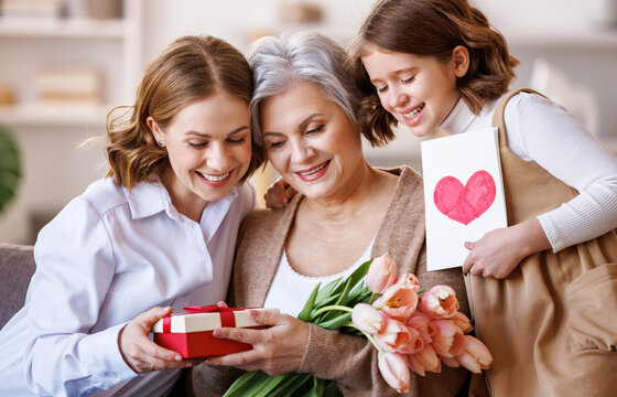 Happy International Women's Day.Smiling  daughter and granddaughter giving flowers  and gift to grandmother   celebrate spring holiday Mother's Day at home