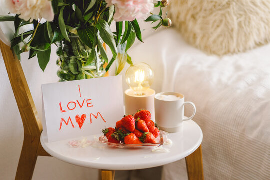 Breakfast for Mothers Day. Heart shaped white plate with fresh strawberries, cup of coffee, gift and Peonys bouquet with gift in bed. Still life composition. Happy Mother's Day.