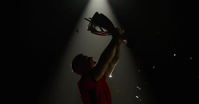 Silhouette of Caucasian male rugby player raising a trophy above head against bright light and falling confetti. Super slow motion, shot on RED cinema camera