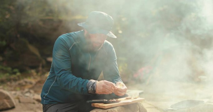 A man cooks food outdoors on a camping trip while sitting by a campfire. Cooking food on a tourist trip to the mountains. Camping and outdoor cooking.