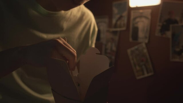 Close-up shot of hands and face of Chinese person eating noodles from takeaway box with chopsticks, at night after working late, photos on wall in background