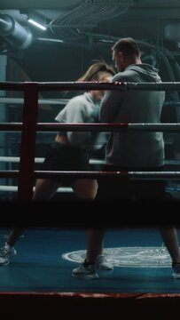 Female fighter in boxing gloves practices fighting technique with male trainer and exercises before championship in dark gym. Boxer hits punching mitts on boxing ring. Vertical shot