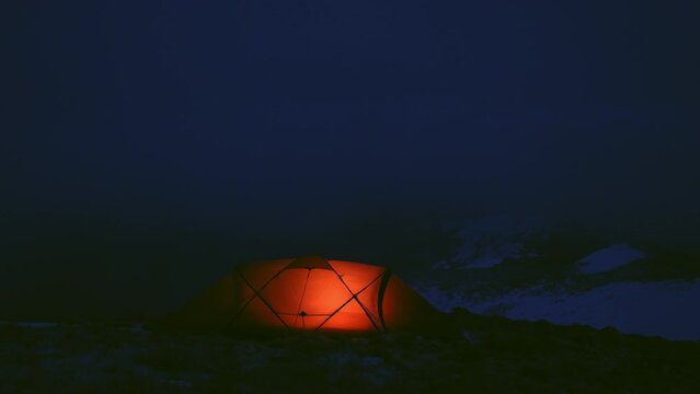 A glowing tent provides warmth and shelter for a man camping in a snowy mountain range under a dark night sky.