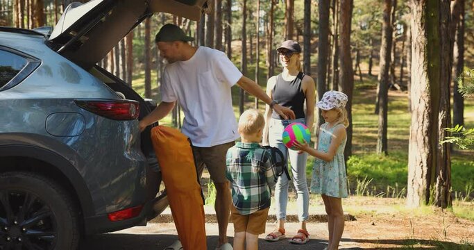 family with kids remove the camping supplies from car trunk at forest. summer vacation, road trip holidays