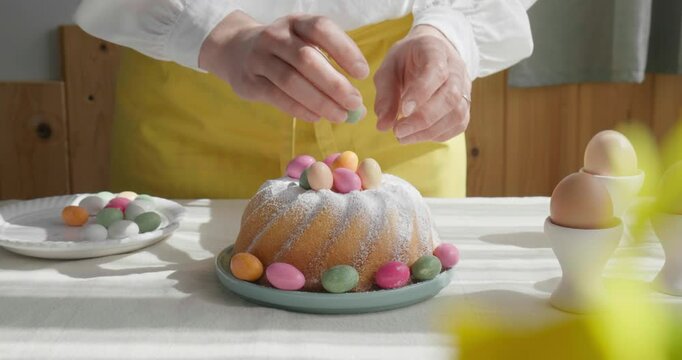 Female hands decorating Easter cake with small colorful chocolate eggs and daisy flowers. Anonymous woman making traditional easter cake or sweet bread with topping. Easter treat.