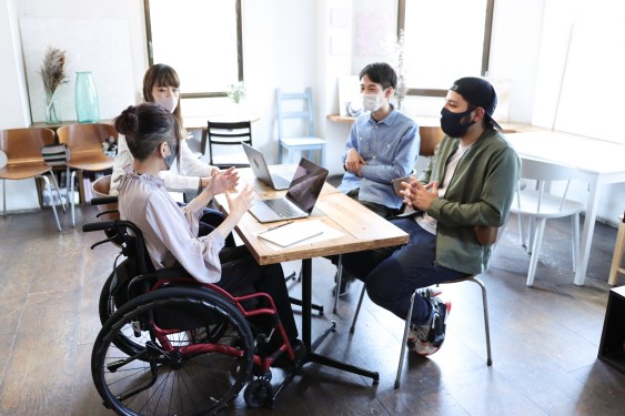 Image of a masked woman in a wheelchair sitting with three masked colleagues at an office.