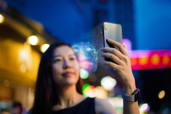 Young Asian woman using face recognition software via smartphone, in front of colourful neon signboards in busy downtown city street at night. Biometric verification and artificial intelligence concept