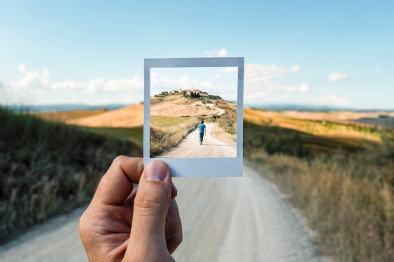 Hand holding up a rectangle with an opening in it to bring something in the distance, a man walking down a dirt road into focus.