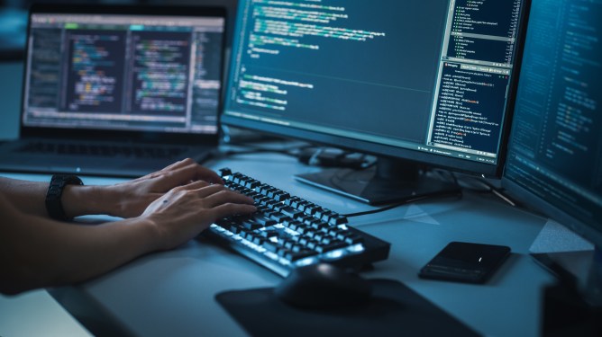 Close up of hands typing code on a keyboard with code appearing on monitor in front of the keyboard.