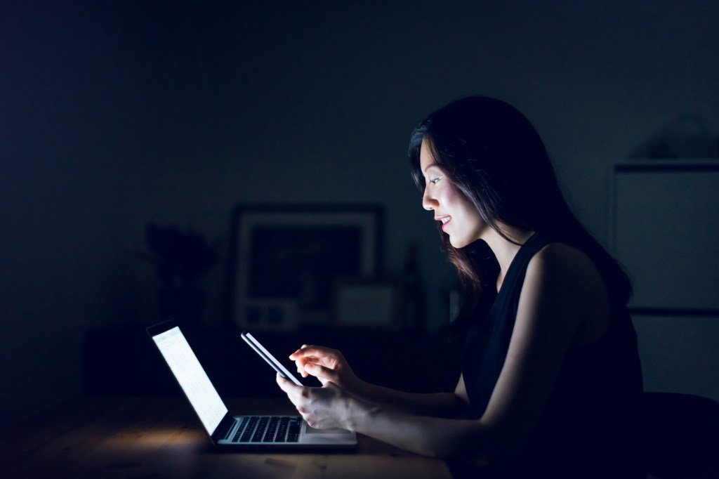 Busy young woman text messaging on smartphone while working on laptop till late in home office, used in a post about mental health app Thoughtfull